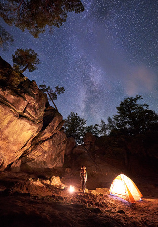 Campsite at night amid huge rock formations. Athletic slim barefooted girl standing straight between small tourist tent and burning bonfire under clear dark starry sky. Tourism and camping concept.の写真素材