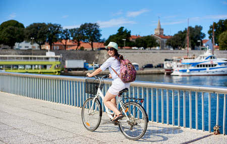 Pretty smiling girl in sunglasses with backpack riding bicycle along fenced stony sidewalk by sheltered harbor of resort town with cruise motorboats. Tourism, active lifestyle and vacations concept.の写真素材