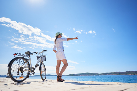 Attractive woman in white closing and sunhat standing at bicycle on stony sidewalk under clear blue sky pointing at opposite shore on sparkling clear water background. Tourism and vacations concept.の写真素材