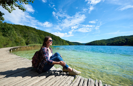 Young smiling tourist woman in sunglasses and with backpack sitting on wooden bridge, enjoying beautiful view, colorful spring panorama of lit by sun green forest and blue lake under clear skyの写真素材
