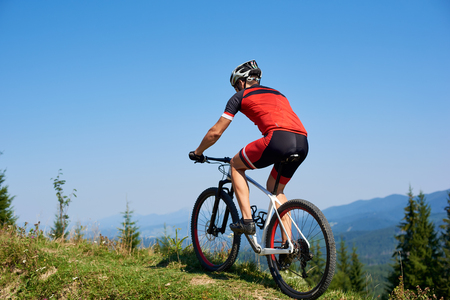 Strong man tourist cyclist in helmet, sunglasses and full equipment riding bike on grassy hill. Mountains and blue summer sky on background. Active lifestyle and extreme sport conceptの写真素材