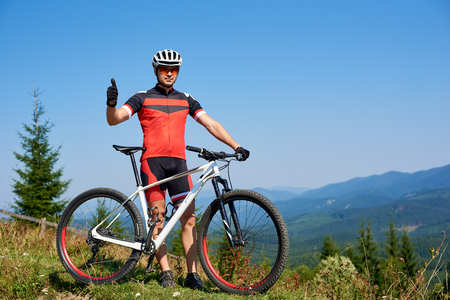 Young athletic successful tourist biker in professional sportswear standing at bike with thumb-up gesture on bright beautiful mountains summer background. Active lifestyle and extreme sport concept.の写真素材
