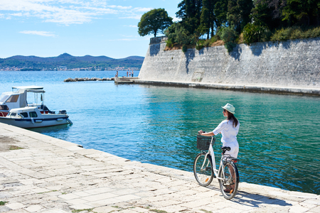 Perfect summer resort landscape. Back view of young woman in sunglasses with backpack riding bicycle along sidewalk by sheltered harbor of resort town with high stone walls. Active lifestyle concept.の写真素材