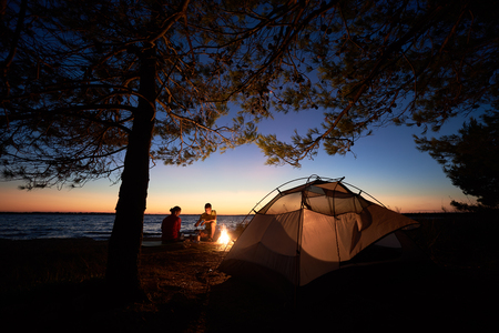 Camping on sea shore at night. Tourist tent under trees and young pair, man and woman preparing food on gas burner near campfire on blue evening sky and water background. Tourism and adventure conceptの写真素材