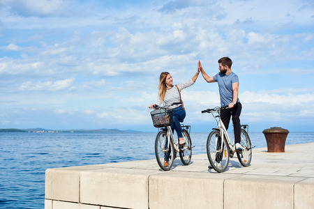 Tourist couple, man and woman standing with bicycles on high paved stone sidewalk, giving high five each other on bright sunny day. Clear blue sea water on background. Active holiday concept.の写真素材