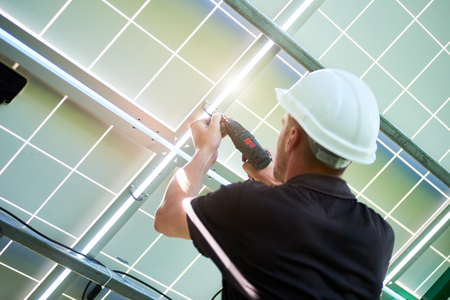 Mounter wearing white helmet and black uniform working with drill installing solar panels. Providing innovative technologies. Modern solution for natural resources saving, using renewable energy.の写真素材