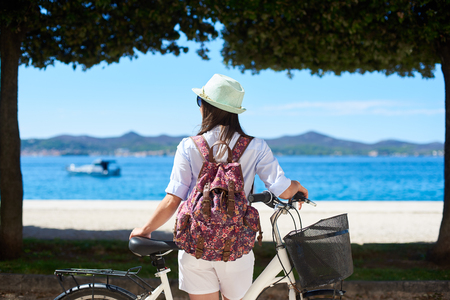 Back view female tourist in white closing, sunhat and backpack standing with bicycle on sidewalk near coast under trees and mountains view on opposite shore background. Tourism and vacations concept.の写真素材