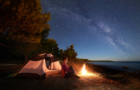 Night camping on sea shore. Smiling woman hiker sitting relaxed in front of tourist tent at campfire under amazing sky full of stars and Milky way. Clear blue water and green forest on backgroundの写真素材