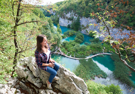 Aeirial view young happy woman sitting on stone, enjoying beautiful view of colorful spring panorama of lit by sun green forest and Plitvice lakes, long wooden bridge with tourists on backgroundの写真素材