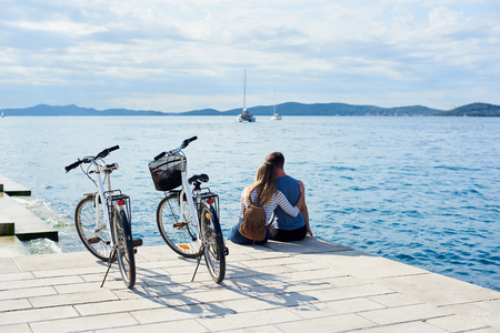 Back view of tourist couple, man and girl with backpack sitting embraced at bicycles on high paved stone sidewalk watching floating cruise yachts in clear blue sea water. Active holiday concept.の写真素材