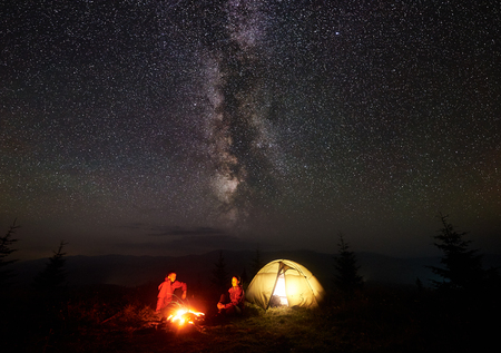 Romantic couple hikers man and woman having a rest on grassy valley near illuminated tent, burning campfire under starry sky and Milky way, enjoying quiet night camping in mountainsの写真素材