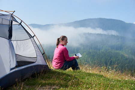Back view of young smiling tourist woman sitting on green grass of blooming valley at tourist tent, reading a book on bright summer morning on foggy mountains background.の写真素材