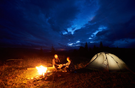 Happy couple man and woman travellers having a rest at night camping in the mountains, sitting near burning campfire and illuminated tourist tent under beautiful evening cloudy sky. Tourism conceptの写真素材