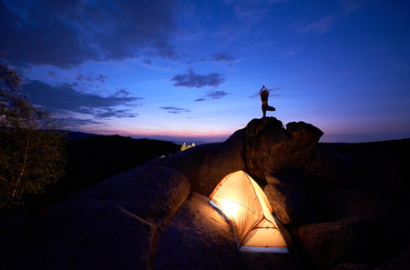 Camping at night on rock formation. Brightly lit tourist tent and small silhouette of woman doing yoga on mountain top against dramatic blue sky at sunset. Outdoor sport, yoga concept. Vrikshasanaの写真素材