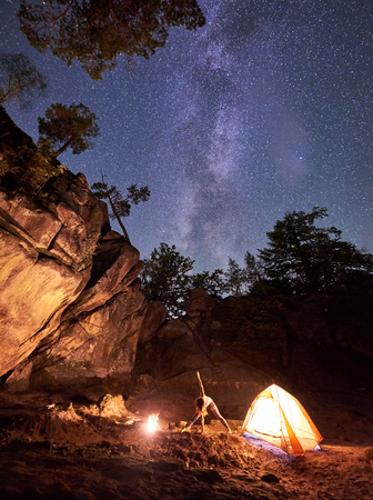 Campsite at night amid huge steep rock formations. Athletic slim girl doing stretching gymnastic exercises at small tourist tent on clear dark starry sky background. Tourism, sport and yoga concept.の写真素材