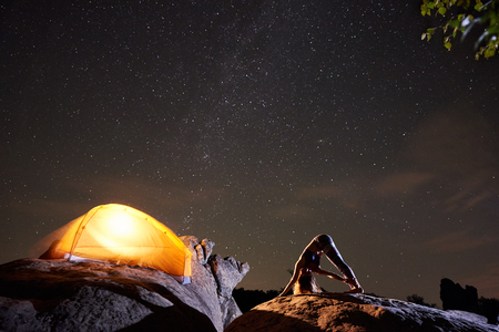Attractive athletic girl doing complicated gymnastic exercises on big boulder on starry sky and brightly lit tourist tent background. Tourism, hiking, active lifestyle, mountain night camping concept.の写真素材