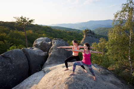 Two slim athletic barefooted girls doing yoga exercises on top of huge rocks on green mountains and clear blue sky background. Climbing, tourism, fitness and healthy lifestyle concept. Virabhadrasanaの写真素材