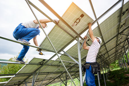 Installing of solar photo voltaic panel system. Two technicians lifting heavy solar module on high platform. Investment in alternative energy, money saving and professional construction concept.の写真素材
