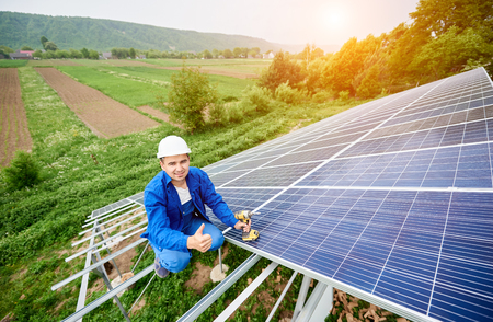Construction worker with screwdriver looking in camera with thumb-up gesture on photo voltaic panel solar system shiny surface and lit by sun green fields background. Alternative energy concept.の写真素材