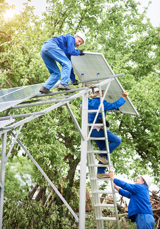 Installing of stand-alone solar photo voltaic panel system. Workers in hard-hats and blue overall lifting the solar module on metal platform. Alternative energy and professional construction concept.の写真素材