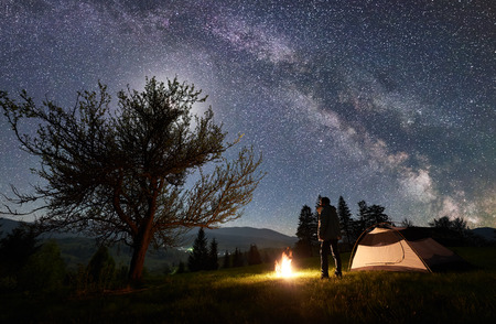 Camping site in mountain valley. Young hiker standing in front of tourist tent at burning campfire near big tree under night blue starry sky with Milky way and moon. Beauty of nature, tourism conceptの写真素材