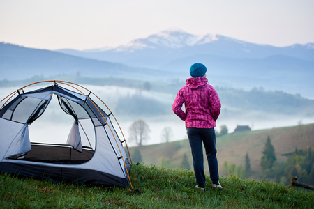Back view of young tourist girl standing near tent enjoying beautiful view of valley covered with dense fog on distant mountains with snowy peaks background. Tourism, camping and recreation concept.の写真素材