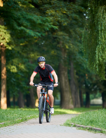 Handsome toned guy bicyclist in cycling clothes, looking to the camera, while riding bike down park alley on summer day. Exercising, reaching goalの写真素材