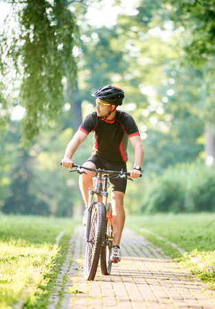 Handsome male biker in professional sportswear looking to side while riding bike down park alley on summer day. Sportsman training thinking about future win in contest. Exercising, reaching goalの写真素材