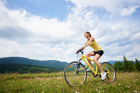 Athlete woman cyclist riding on yellow mountain bicycle on a grassy hill, enjoying summer day in the mountains. Outdoor sport activity, lifestyle conceptの写真素材