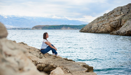 Young attractive female tourist in jeans and T-shirt sitting alone on stony shore of sea, enjoying beautiful view. Clear shiny water and mountains on background. Tourism and vacations concept.の写真素材