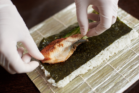 Close-up view of process of preparing rolling sushi. Nori, white rice, cucumber and eel on bamboo mat. Chefs hands in gloves starts cooking sushi rollsの写真素材