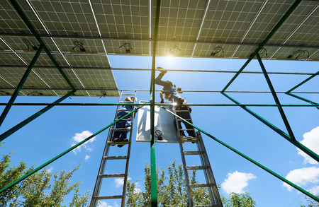 Professional workers team installing solar panels on the green metal construction using different equipment. Innovative solution for energy solving. Use renewable resources. Green energy.の写真素材