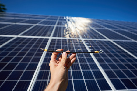 cropped photo of man's hand keeping part of red wirs needing by solar panels installing. Huge solar battery on glancing with sunblinds on background. Modern solution for natural resources saving.の写真素材