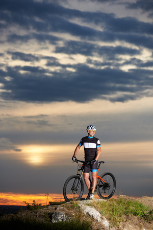 View of handsome and robust man smiling and standing near bicycle on rock hill. Positive cyclist holding his hands on handle bar of bike and posing. Concept of healthy lifestyle, sports and motivationの写真素材