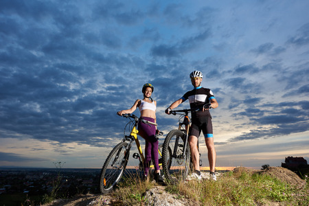 Two positive cyclists holding hands on handle bars of their bikes, looking at camera and smiling. Beautiful girl wearing violet leggings and white top, man in black sportswear and sport glasses.の写真素材