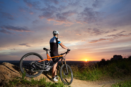 Back view of cyclist male in a helmet and sports clothes rests on the background of a sunset from a height watching the mountain in the distance, the enchanting sky and the bright sunの写真素材