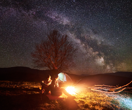 Camping night in mountains. Dark silhouettes of two sitting young tourists, boy and girl lit by burning campfire under dark starry sky on tent and distant hills background. Tourism and travel concept.の写真素材