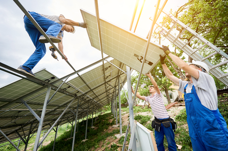 Stand-alone solar voltaic panel system installing. View from below of three technicians workers mounting heavy solar panel. Alternative energy production and professional construction concept.の写真素材