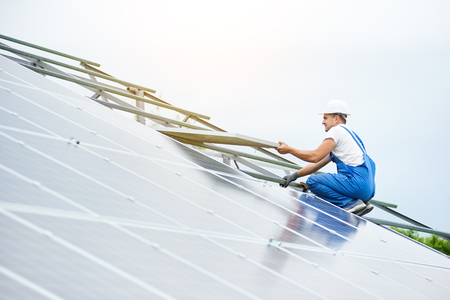 Installing of solar  voltaic panel system. Professional worker in hard-hat and blue overall lifting the solar module on metal platform. Alternative energy and professional construction concept.の写真素材