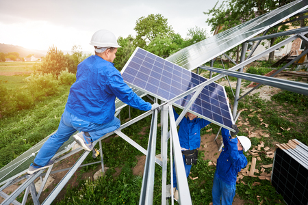 Installing of stand-alone solar voltaic panel system. Team of three technicians in hard-hats mounting the solar module on platform on green summer view background. Alternative energy concept.の写真素材