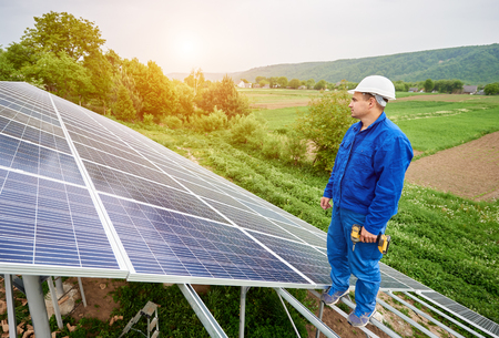 Construction worker with screwdriver standing on metal frame of voltaic solar system looking on shiny surface. Alternative energy, ecology protection and cheap electricity production concept.の写真素材