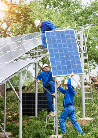 Installing of stand-alone solar voltaic panel system. Workers in hard-hats and blue overall lifting the solar module on metal platform. Alternative energy and professional construction concept.の写真素材