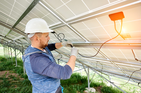 Installing and wiring of stand-alone solar photo voltaic panel system. Close-up of young electrician in hard-hat connecting electrical cables inside the solar modules. Alternative energy concept.の写真素材