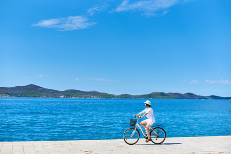 Beautiful female cyclist in white summer closing and hat riding bicycle along stony sidewalk on blue sea water and resort town at foot of mountains on opposite shore background. Tourism and vacations.の写真素材