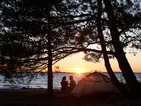 Back view of silhouettes of young tourist couple man and woman sitting on lake shore in front of small tent on blue clear water background enjoying beautiful sunset. Tourism and camping concept.の写真素材