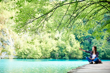 Young happy tourist woman with backpack sitting on wooden bridge enjoying beautiful view of colorful spring panorama of lit by sun green forest, pointing at distanceの写真素材