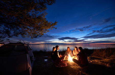 Night summer camping on sea shore. Group of five young tourists sitting on the beach around campfire near tent under beautiful blue evening sky. Tourism, friendship and beauty of nature concept.の写真素材