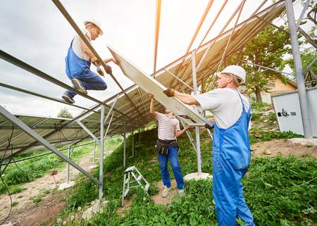 Installing of solar photo voltaic panel system. Three technicians lifting heavy solar module on high platform. Investment in alternative energy, money saving and professional construction concept.の写真素材
