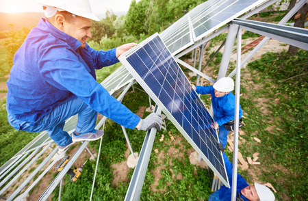 Team of three technicians mounting photo voltaic panel to stand-alone solar system platform on bright sunny day. Alternative energy, professionally done job and financial investment concept.の写真素材