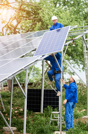 Installing of stand-alone solar photo voltaic panel system. Workers in hard-hats and blue overall lifting the solar module on metal platform. Alternative energy and professional construction concept.の写真素材